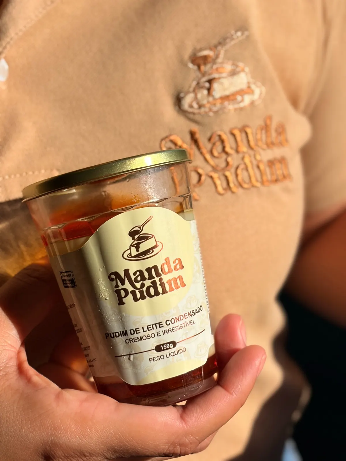 Mão segurando o pudim 150g pronto pra servir no clássico copo americano com o rótulo da Manda Pudim